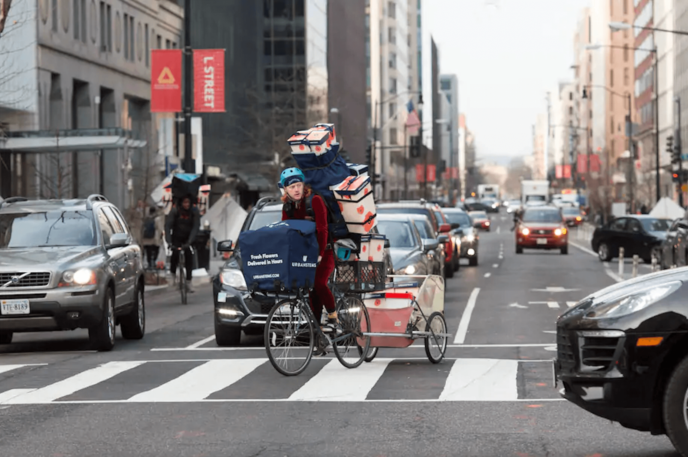 Courier crossing a NYC intersection with a tower of UrbanStems flower boxes stacked on her bike